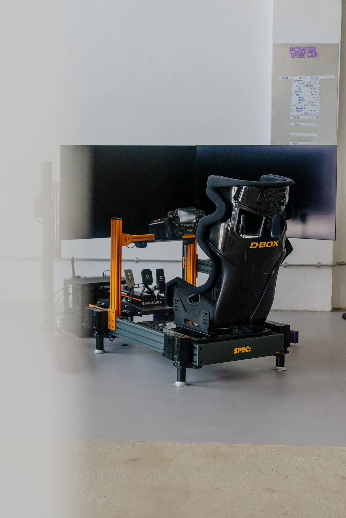 A SPEC 282 sim rig with orange uprights and a motion seat, set up in a studio in front of triple screens.