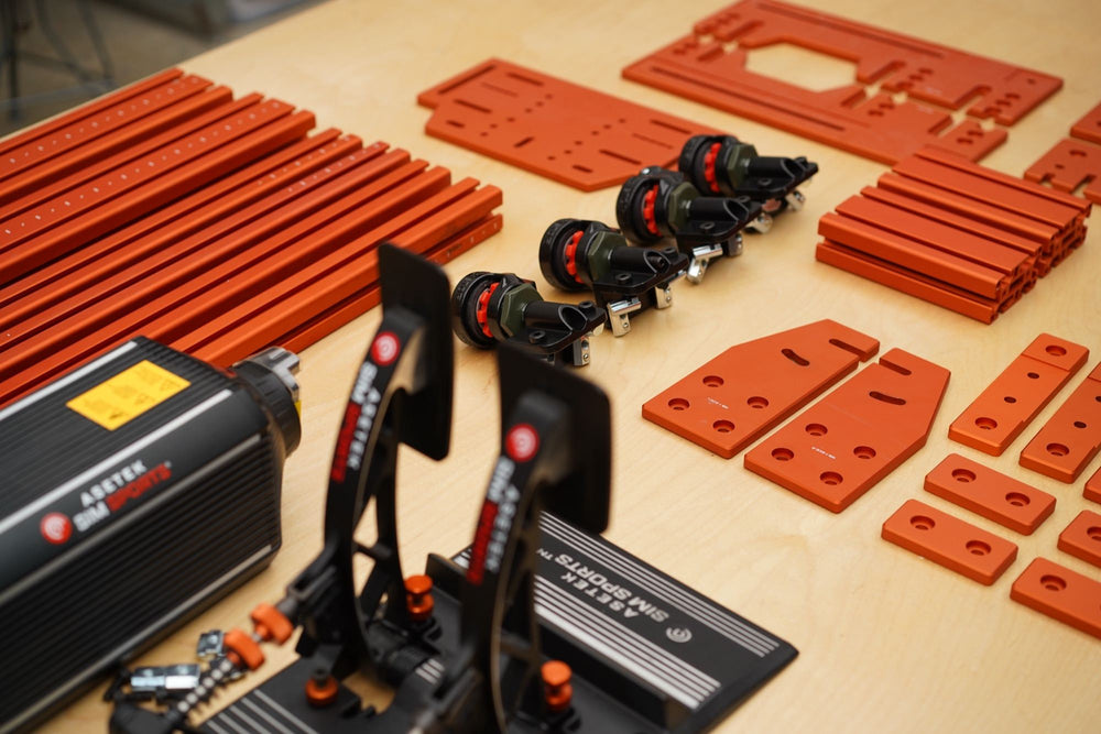 Orange SPEC 282 parts laid out on a bench beside an Asetek wheelbase, pedals and casters