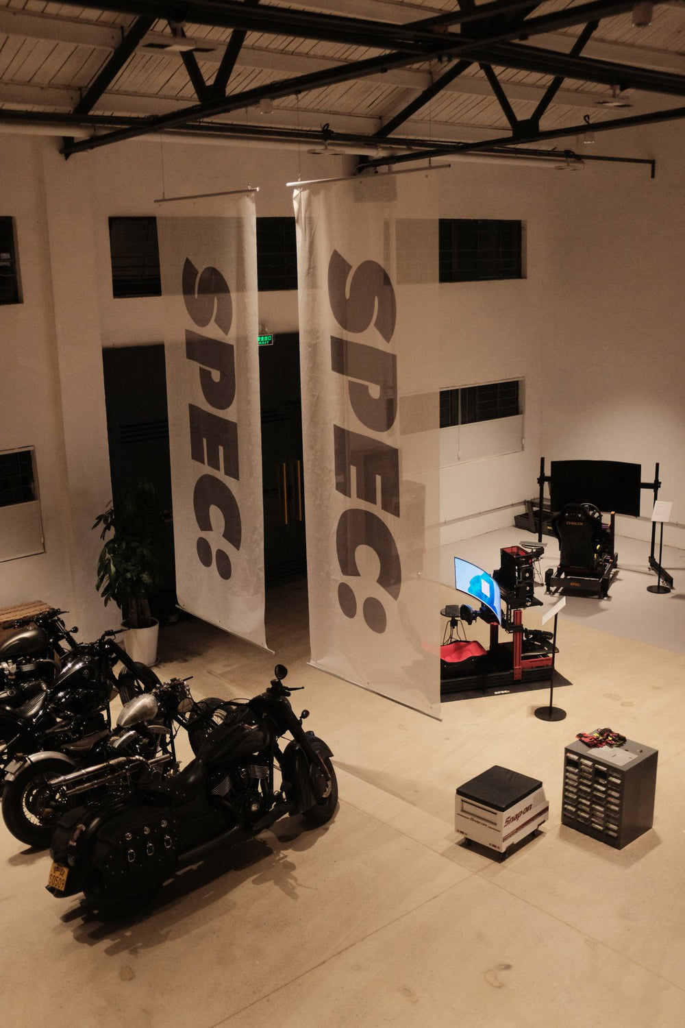 SPEC banners hanging from the studio roof trusses, a sim rig and motorcycles on the floor below